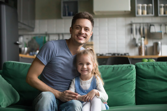 Portrait of happy smiling father with preschool daughter, father sitting on sofa at home together with little girl, kid sitting near dad, look at camera