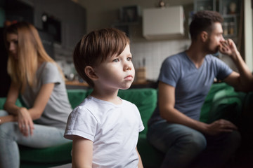 Upset frustrated little boy tired of parents fight, toddler son looking out window reaming that family conflicts would stop while parents sitting separately on sofa, suffering from mom and dad quarrel