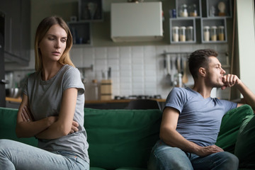 Offended upset man and woman sitting separately on couch, sofa in living room, wife looking with...