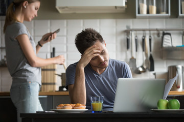 Young couple using laptop and smartphone in the kitchen, upset man working with documents, reading...