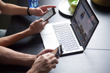 Young couple using smartphones with laptop, texting, checking social networks at home, female and male hands holding phones, browsing websites concept, close up