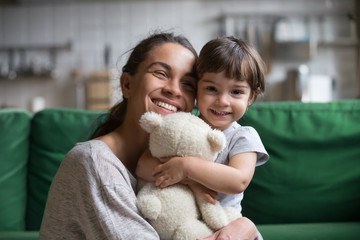 Smiling single young mum embracing little preschool daughter with toy, playing in living room at home, mother laughing with child, headshot portrait, cute girl look at camera