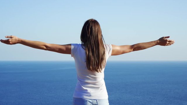 Young woman in white t-shirt raising arms up at cliff against breathtaking view of blue Mediterranean sea. Carefree female outstretching hands. Concept of freedom and inspiration
