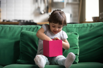Excited curious little preschool cute girl opening, checking gift box, sitting on sofa, couch,...