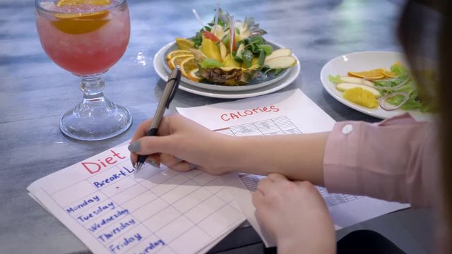 nutritionist woman writes diet plan with food calorie sheet on table with punch in glass and fruits in plate at cafe