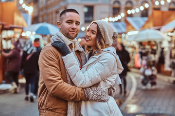 Fototapeta premium A young romantic couple wearing warm clothes hugging outdoor in evening street at Christmas time, enjoying spending time together.