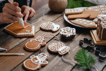 A young girl decorates ginger cookies Christmas winter morning. Woman draws Icing on honey gingerbread house. Wooden brown table. copy space. Blank biscuit gingerbread house, ready to decorate.