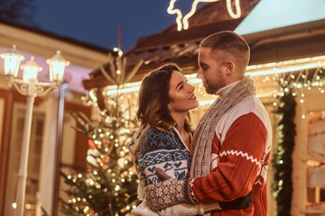 A young romantic couple wearing warm clothes hugging outdoor at Christmas time, standing in evening street decorated with beautiful lights.