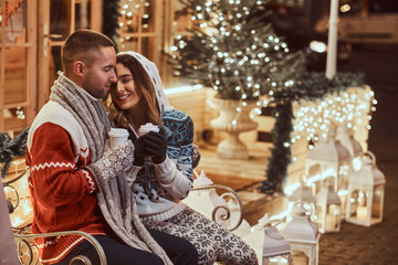 A young romantic couple wearing warm clothes outdoor at Christmas time, sitting on a bench and warming with hot coffee in evening street decorated with beautiful lights.