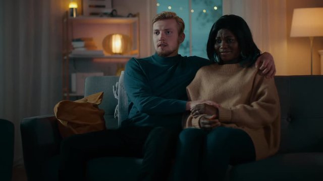 Happy Young Couple Watching Horror Movie on TV while Sitting on a Couch, they got Scared, Girl Covers eyes. Handsome Caucasian Boy and Black Girl in Love Spending Time Together. Moving Camera Shot.