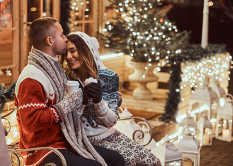 Fototapeta na wymiar A young romantic couple wearing warm clothes outdoor at Christmas time, sitting on a bench and warming with hot coffee in evening street decorated with beautiful lights.