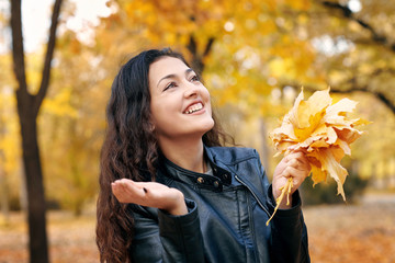 Pretty woman is posing with bunch of maple's leaves in autumn park. Beautiful landscape at fall season.