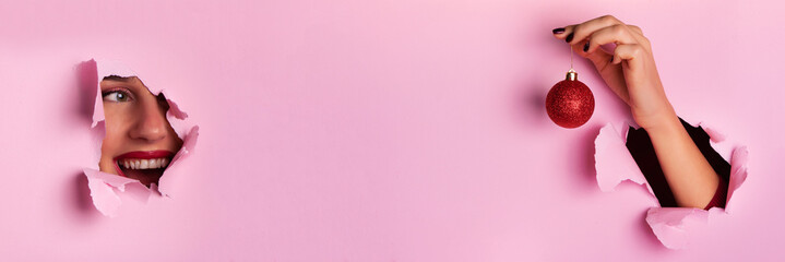 New year celebration banner, copy space. Surprised face in paper hole with metalic effect, hand holding red glittering Christmas ball on pink background.