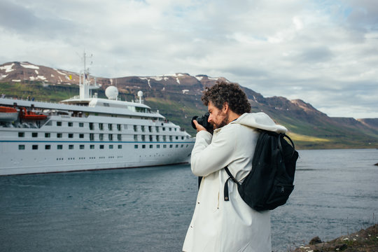 Trendy, millennial photographer in white raincoat or jacket and black hipster backpack makes photo of white cruise ship leaving port. New trend in tourism and travelling, explore iceland and norway