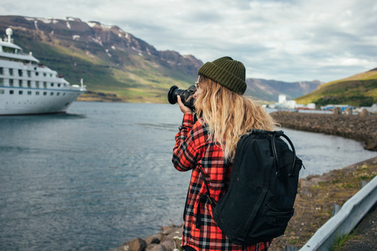 Young trndy millennial hipster, blonde woman in red shirt and green beanie makes photos of huge cruise ship entering fjords in iceland or norway. Travel blogger explores world