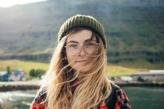 Portrait of young natural beautiful woman or girl with bright blue eyes and blonde hair in wind, wear green beanie and red hipster shirt, look at camera, gently smile