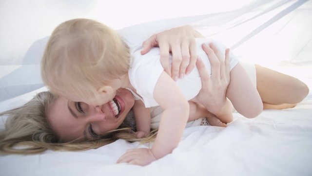 Cute baby boy playing with loving mother relaxing under the white scarf in the park 