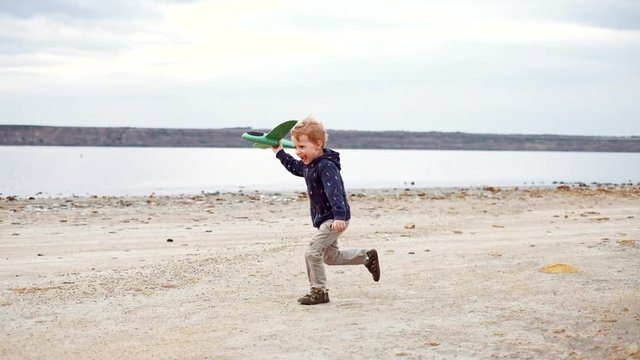 Happy child running with toy airplane and launching it in the sky. Kid boy playing with plane outdoors on nature. Little pilot dreaming of flight, having fun on vacation with family. Slow motion.