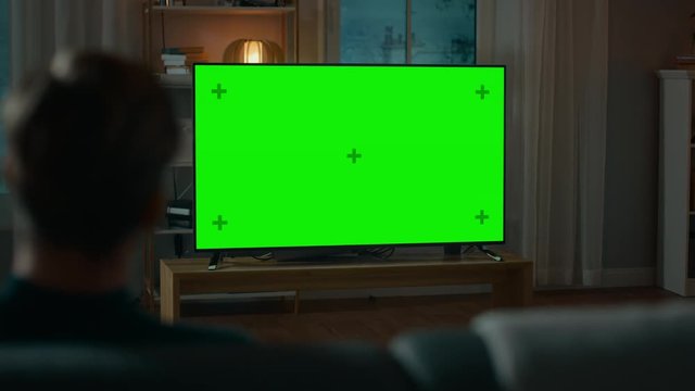 Man Watches Green Mock-up Screen TV while Sitting on a Couch at Home in the Evening. Cozy Living Room with Warm Lights. Over the Shoulder Shot.