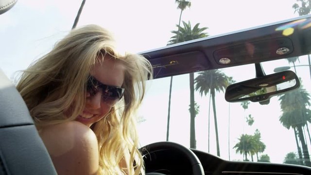 Young blonde American girl enjoying summer sunshine driving luxury cabriolet along palm tree lined suburban street 