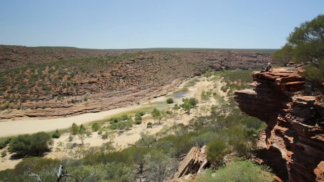Caucasian woman resting on red sandstone rock along the trail to Nature's Window over the Murchison River in Kalbarri National Park, Western Australia. Blonde backpaker enjoying in Australian outback.