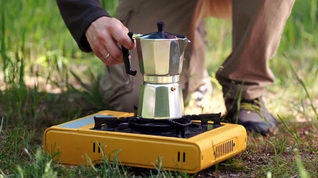 Traveler hands lights camping stove, holding coffeepot and pouring coffee in cup. Tourist man cooking with coffee maker and drinking from mug on picnic. Hiker with travel pot outdoors on nature.