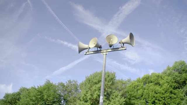 Outdoor public address loudspeakers against a blue sky and trees in slow motion