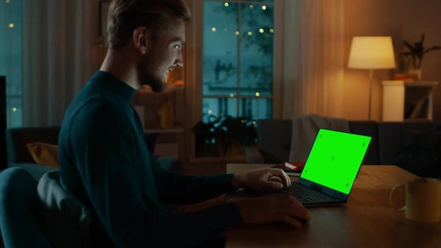 Man Sitting at His Desk Works on a Laptop with Green Mock-up Screen. Late at Night in His Living Room Man Uses Notebook Computer. Side View Zooming on Screen.