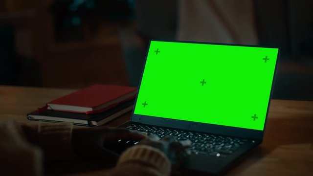 Beautiful Black Woman Sitting at Her Desk Works on a Laptop with Green Chroma Key Screen. Late at Night in Her Living Room Girl Uses Notebook Computer. Over the Shoulder Shot.