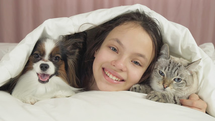 Happy teen girl communicates with dog Papillon and Thai cat in bed