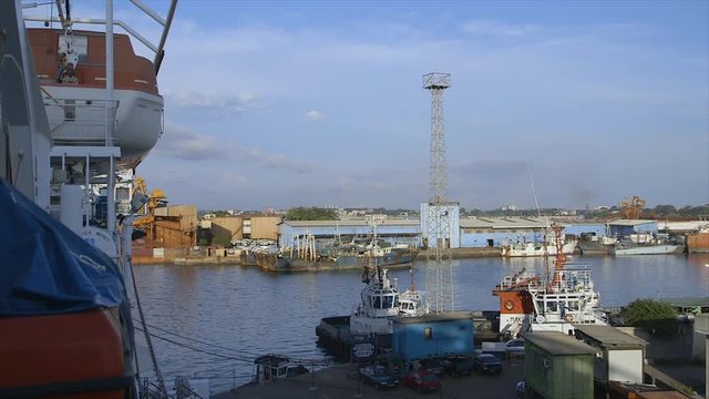 Wide, clear, still shot of ships docking area, section of Mercy Africa dwarfing all smaller ships and ferries,  calm clean water, long storage building, blue sky with low grey clouds, flood ligh