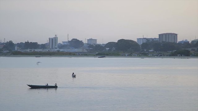 Wide, still shot, light grey sky, serene water, sunset surface reflections, birds fly, partial silhouettes, two fishermen paddling their canoe towards shore, misty horizon land, trees, buildings