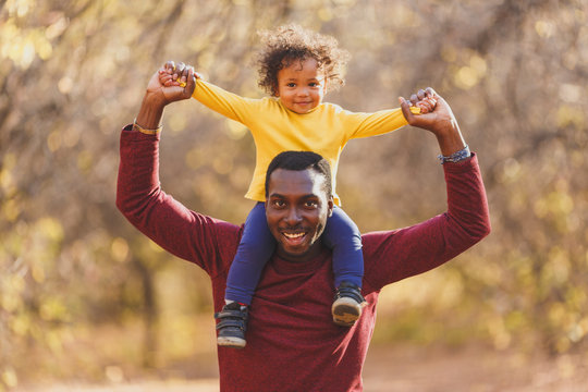 Smiling Father Holding His Son On Shoulder And Looking At Camera. Portrait Of Happy African Dad Little Boy Smiling In The Garden.