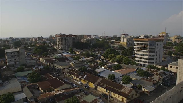 Wide still aerial shot of Conakry city street, Guinea, morning sun-rays, close buildings, quiet road, rusty galvanised steel roof houses, few  flats, lash tall trees, telecommunication and light pylon