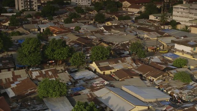 Medium aerial still shot, morning view, partial sun rays,  rusty galvanized  iron sheet roofs, congested slum houses on either side of the street, road, stones, plastic covers on roof-tops,  poor acc