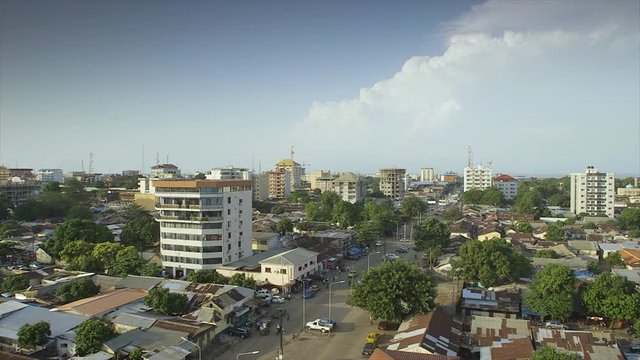Wide still shot,  aerial view, calm morning,  Conakry city highway road, clean,  streetlights, low traffic, people walking, leafy trees,  rusty roofs,  few modern flats, white blue clouds, ho