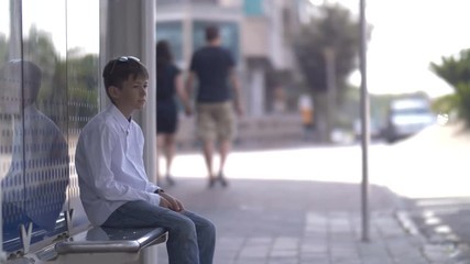 boy sits on the bus stop and waits for the bus, looks at watch