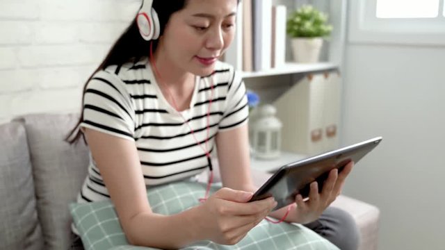 from down to up asian lady student relaxing sitting on sofa talking by earphones on tablet online learning. young girl talking with tutor on internet in bright living room sunshine from window.