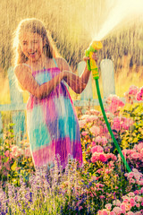 Playfull girl watering flowers with rain in the garden at summer day. Child using garden hose on...