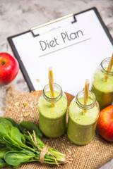 The diet plan is written on a white sheet on the tablet among the bottles of green smoothies, spinach and apples on a gray table with burlap. Weight loss concept. Top view