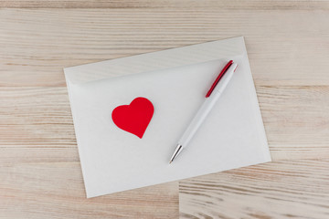 любовная почта:  белых пустых незаполненных конверт для писем, белая шариковая ручка и красное сердечко  лежит сверху,  светлый деревянный стол