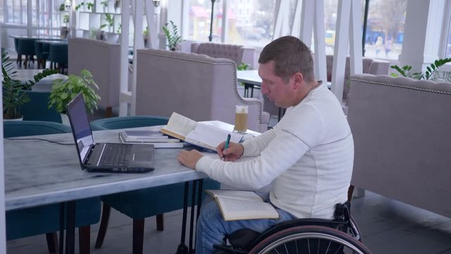 distance education of invalid, successful sick student male on wheelchair working with laptop to learn from online lessons and making notes in notebook in cafe