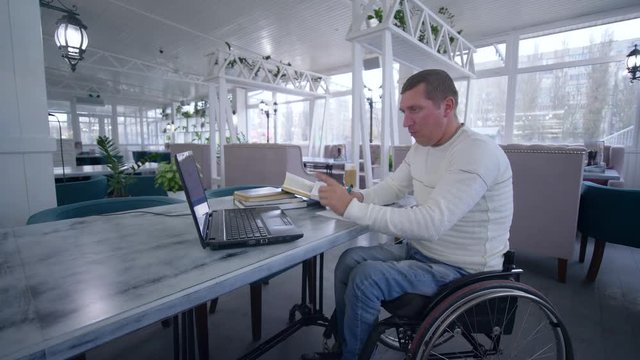 online education of handicapped, student disabled man on wheelchair uses modern laptop technology to learn from online lessons and books making notes in notebook close-up sitting at table in cafe