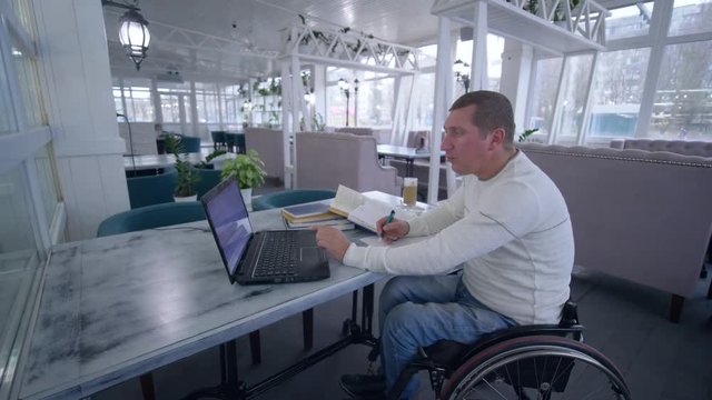 online education, successful student invalid men on wheelchair uses modern laptop technology to learn from online teaching and books making notes in notebook close-up sitting at table in cafe