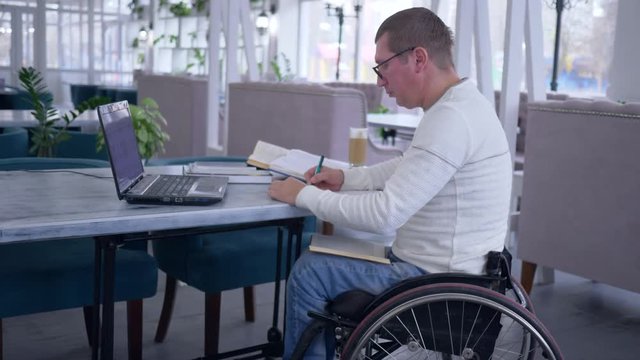 online teaching, successful aching man on wheelchair is learning for self-development uses modern technology for distance education and makes notes in notebook in restaurant