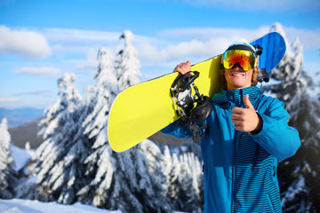 Smiling snowboarder posing carrying snowboard on shoulders at ski resort near forest before freeride session. Rider showing thumb up sign wearing polarized goggles. Modern snowboarding equipment.