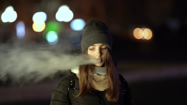 A girl is smoking vape outside in the evening. Nice colorful bokeh in front of the evening city.
