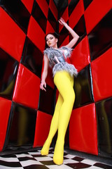 crazy fashion girl wearing yellow pantyhose, silver furwaistcoat and futuristic steel belt posing near red wall with black squares alone
