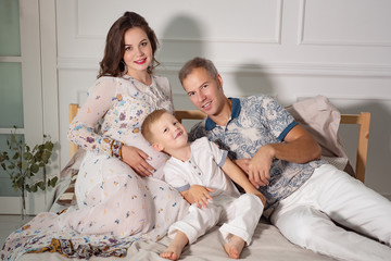 Adult man with pregnant woman and little boy sitting on bed cuddling against white background