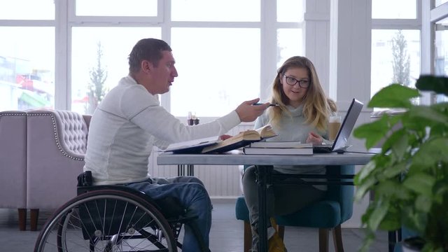 individual studying for disabled, teacher woman into eyeglasses conducts lecture for invalid male on wheelchair using a laptop computer and books in cafe